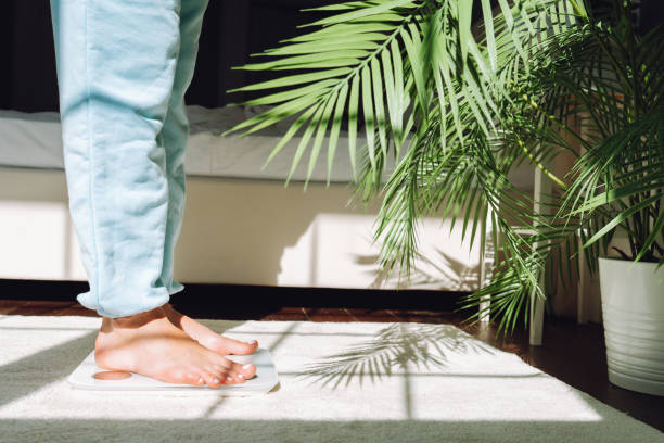 unrecognizable woman control her weight, standing on scales near plant indoors